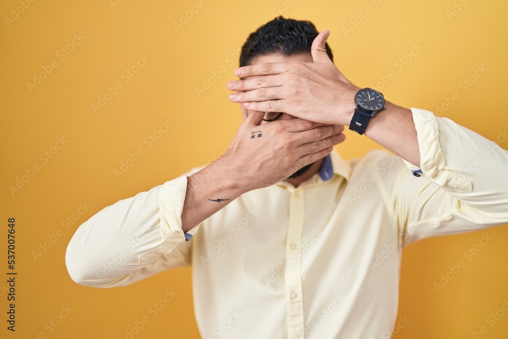 Sticker Hispanic young man wearing business clothes and glasses covering eyes and mouth with hands, surprised and shocked. hiding emotion