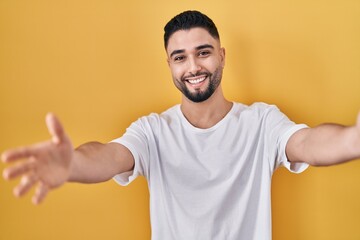 Young handsome man wearing casual t shirt over yellow background looking at the camera smiling with open arms for hug. cheerful expression embracing happiness.