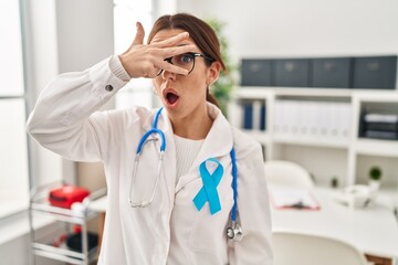 Young brunette doctor woman wearing stethoscope at the clinic peeking in shock covering face and eyes with hand, looking through fingers with embarrassed expression.