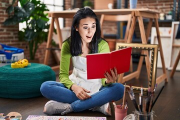 Young caucasian woman artist sitting on floor reading book with surprise expression at art studio