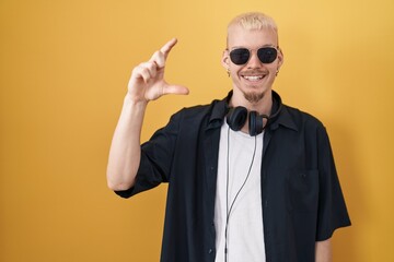 Young caucasian man wearing sunglasses standing over yellow background smiling and confident gesturing with hand doing small size sign with fingers looking and the camera. measure concept.