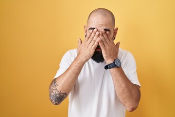 Young hispanic man with beard and tattoos standing over yellow background rubbing eyes for fatigue...