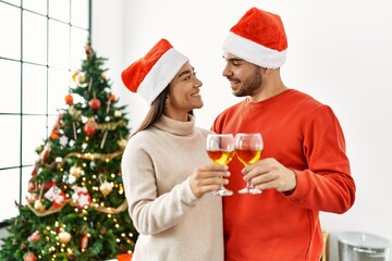 Young hispanic couple smiling happy toasting with champagne standing by christmas tree at home.