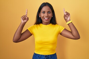 Young indian woman standing over yellow background smiling amazed and surprised and pointing up with fingers and raised arms.