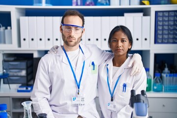 Interracial couple working at scientist laboratory thinking attitude and sober expression looking self confident