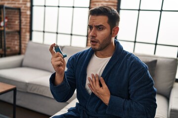 Young hispanic man using inhaler sitting on sofa at home