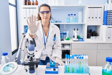 Young hispanic woman working at scientist laboratory doing stop sing with palm of the hand. warning...