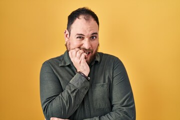 Plus size hispanic man with beard standing over yellow background looking stressed and nervous with hands on mouth biting nails. anxiety problem.