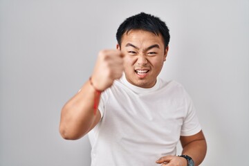 Young chinese man standing over white background angry and mad raising fist frustrated and furious...