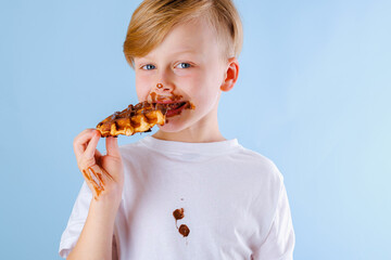 The child is eating Belgian waffle with chocolate sauce on a blue background. Dirty chocolate...