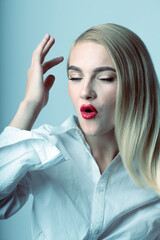 Fashion and make-up concept. Close-up studio portrait of beautiful blonde woman with red lipstick and closed eyes making O shape with lips. Model wearing white shirt. Toned image with blue color