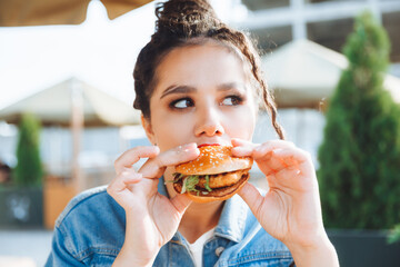 A young glamorous woman with dreadlocks and red lipstick is sitting and eating a burger in a street...
