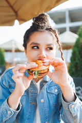 A young glamorous woman with dreadlocks and red lipstick is sitting and eating a burger in a street...