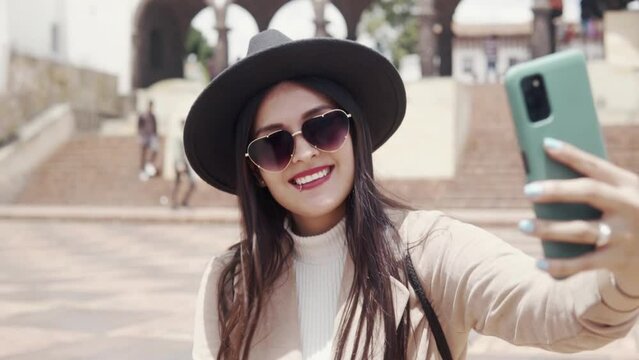 A Latin young adult woman with glasses, coat and hat is smiling and taking a selfie in a square