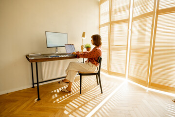 Woman works on computers while sitting by a cozy workplace at sunny living room with blinds on the...