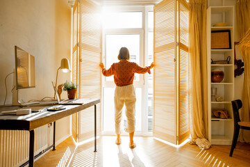 Woman opens window blinds letting the sun inside the room, spending good morning in sunny and cozy apartment in beige tones. Interior view
