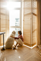 Young woman sits with her dog by the window blinds and looks away in cozy and sunny living room of...