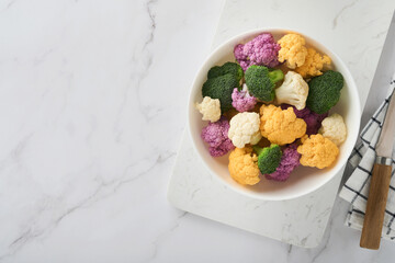 Fototapeta na wymiar Colorfu cauliflower. Cauliflower cut into small pieces white bowl on light stone marble concrete background. Purple, yellow, white and green color cabbages. Agricultural harvest. Mock up. Top view.
