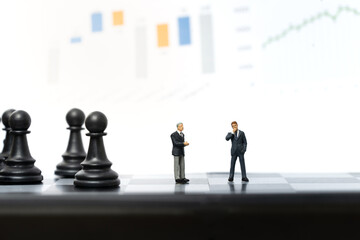 Miniature people, two businessmen stand on chessboard with graph on background. Business and financial concept