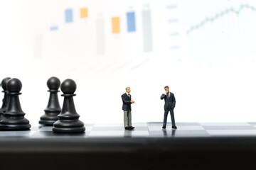 Obraz premium Miniature people, two businessmen stand on chessboard with graph on background. Business and financial concept