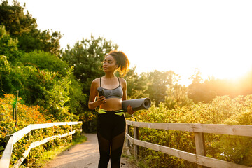 Young black woman walking with yoga mat after practice in summer park