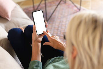 Senior caucasian woman sitting on sofa in living room, using smartphone with copy space