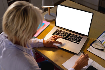 Senior caucasian woman sitting at table in kitchen, using laptop with copy space