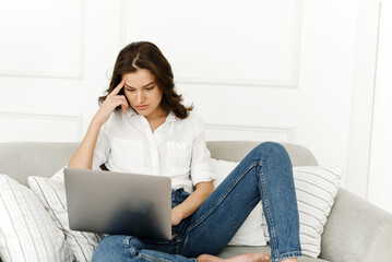 Serious focused young woman using laptop, checking news on internet, sitting on sofa, solving problems at remote work