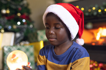Happy african american boy holding christmas snowing ball and siting next fireplace