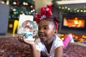 Happy african american girl holding christmas snowing ball and siting next fireplace