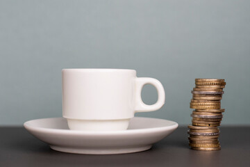 Cup of coffee on a small white plate with euro coins next to it forming a tower on a gray and black...