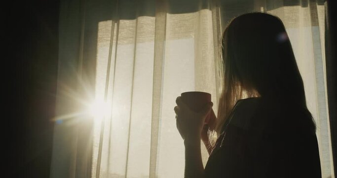 A woman with a cup of hot tea looks out the window where the sun has risen. Hot drink and good mood