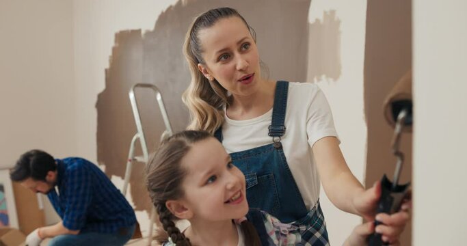 Family is renovating house and painting walls. Mother with curly hair shows daughter with how to properly paint wall. They are holding roller together, talking and smiling. Dad is painting wall behind