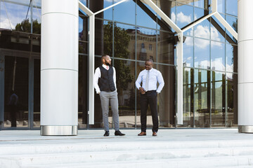 Portrait of two dark-skinned businessmen walking and talking in front of a modern building exterior. Friendly meeting outdoors