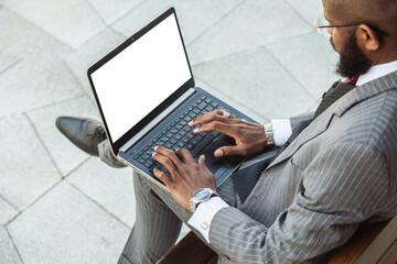 A dark-skinned man in a business suit is using a laptop outdoors. Working break. Online learning and education. Distance courses and communication. Copy space and mock up