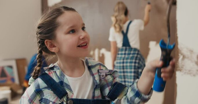 Beautiful seven-year-old girl with pigtails paints wall with roller and smiles. Girl enthusiastically helps parents. She is wearing checkered shirt and denim overalls. Behind, mother is painting wall.