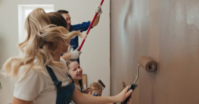 Woman with curly hair in white t-shirt paints wall with family. Husband and two children are holding rollers and painting the wall brown. They dance, smile, have fun and sing loudly.