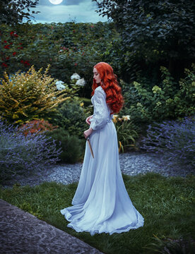 Fantasy photo red-haired woman holding weapon dagger. Long flowing red hair white vintage old style dress hem train. Girl sexy aristocrat back rear view walks in night summer garden green nature trees