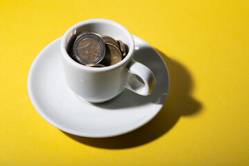 A white color coffee cup with small saucer with euro coins inside the cup on yellow color background