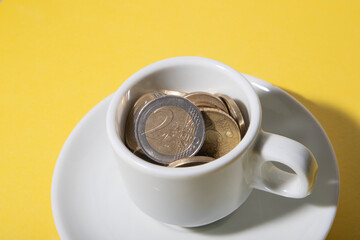 A white color coffee cup with small saucer with euro coins inside the cup on yellow color background