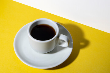 Taza de café de color blanco con plato pequeño de color blanco sobre fondo amarillo.
