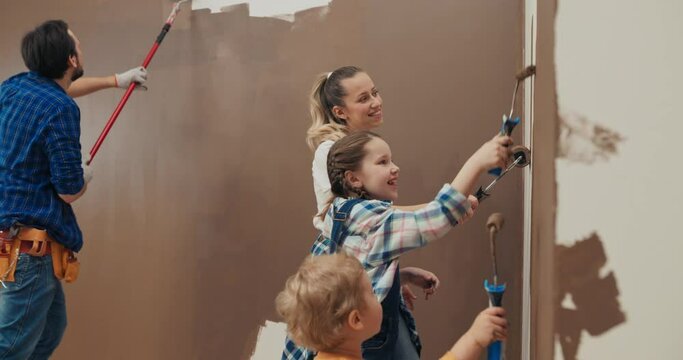 Happy family is painting the walls in new apartment. Parents and children have rollers in hands and paint the wall in brown. Mom and dad and two children sing, dance, smile and have fun.