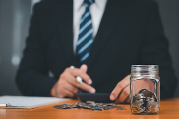 Money in glass jar on background, businessman in suit is using cost calculator with stack of coins in front. Financial concepts, savings, money management, taxes.