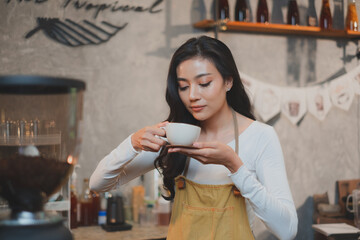 Beautiful Asian female Barista coffee maker or staff is holding  and smelling a cup of coffee with smile and happy emotional in a coffee shop or cafe. Concept of customer service, Service minds.