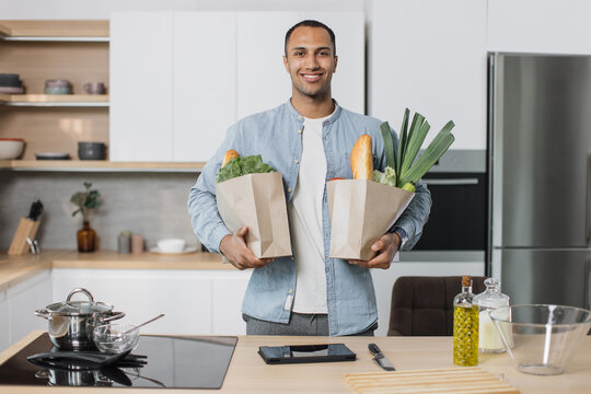 Young Attractive Indian Man In Kitchen Holding Two Paper Grocery Shopping Bags Full Of Fresh Healthy Food Vegetables In His Hand, Standing On The Background Of New Modern Light Kitchen At Home.