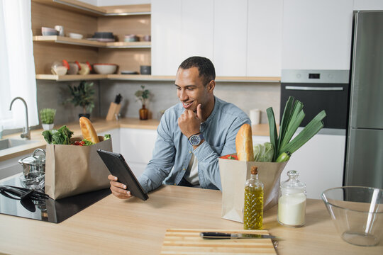 Attractive Young Indian Man Using Digital Tablet Pc For Reading Recipe Of Vegan Vegetable Salad, Sitting And Smiling While Cooking In Modern Bright Kitchen At Home.