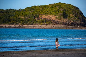 Fototapeta na wymiar A beautiful girl in a long dress stands on the ocean shore with the mighty mountains in the background; a colorful sunset on the beach in cape hillsborough national park, vacation in queensland, austr