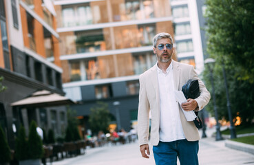 Cheerful middle age businessman in glasses walking by street towards modern office building, going to job in the morning