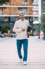 Cheerful middle age businessman walking by street towards modern office building, going to job in the morning