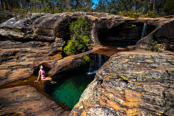 beautiful girl in pink bikini sits by natural rock pool with waterfalls at rainbow waterfall in blackdown tableland, holiday in australian outback, queensland